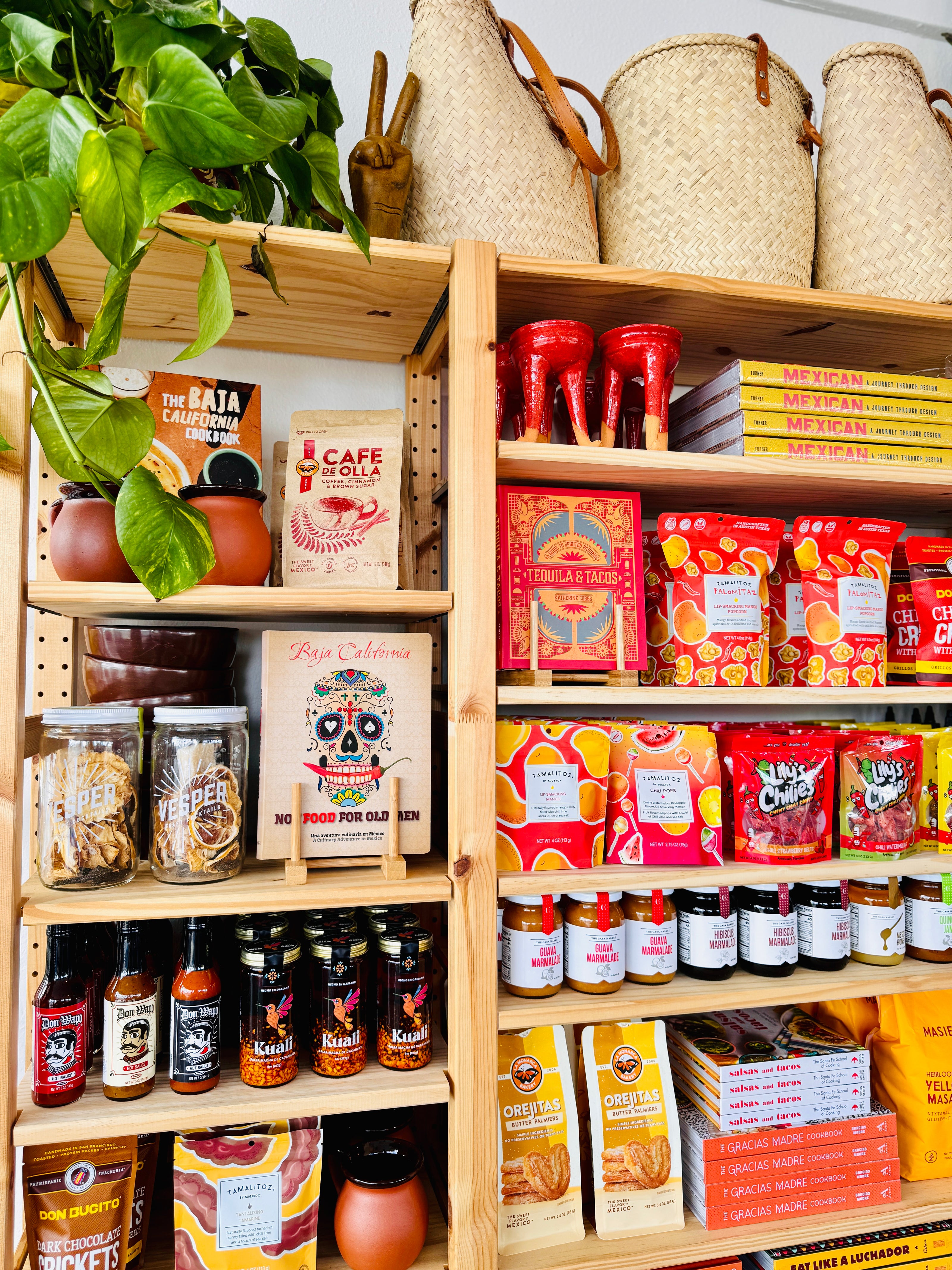 Photo of stocked shelves containing cocktail mixes, cookbooks, Don Wapo hot sauce, Kuali chili oil, dark chocolate covered crickets, tamarind flavored gourmet candy, traditional Mexican barro mug, gourmet popcorn, chili covered gummy candy, cookies, & masa mix